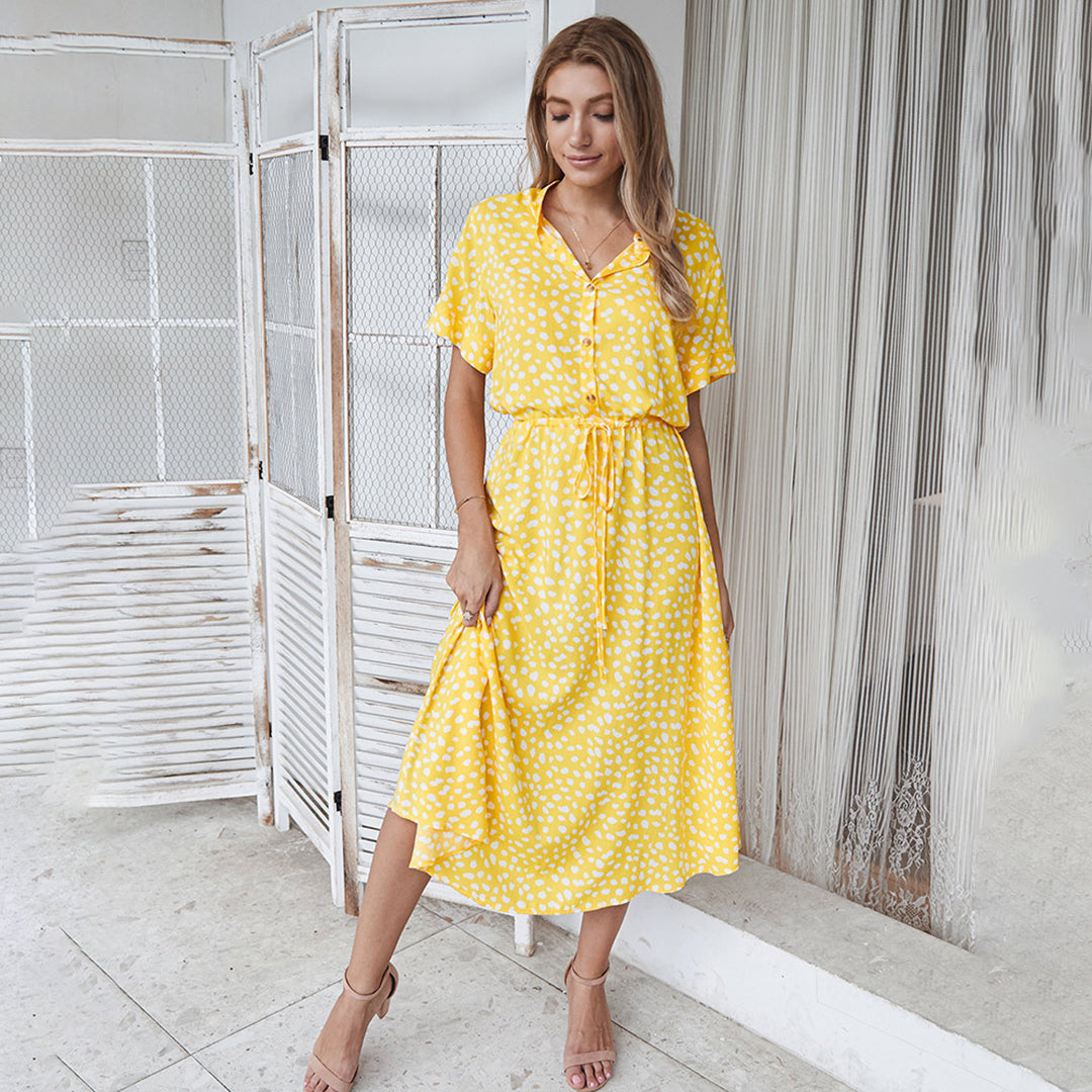 Vrouw in gele zomerjurk met witte stippen, staand voor een witte achtergrond. Modieuze dameskleding, casual stijl, zomermode.