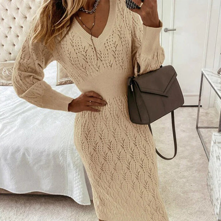 Beige gebreide jurk met V-hals en lange mouwen, gedragen door model in slaapkamer. Stijlvolle damesmode, herfstkleding, elegante handtas.