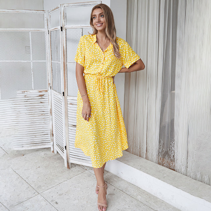 Vrouw in gele zomerjurk met witte stippen, staand voor een witte achtergrond. Modieuze dameskleding, casual stijl, zomermode.