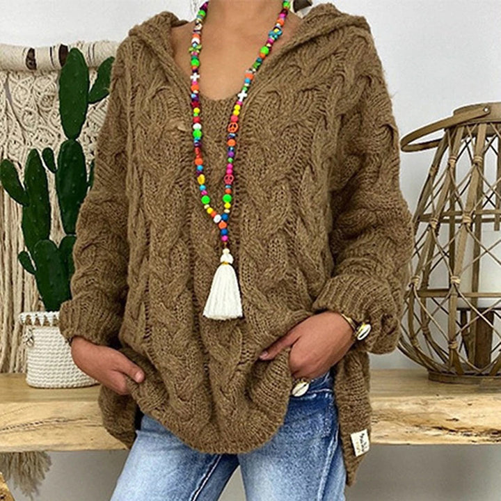 Vrouw draagt bruine gebreide trui met kabelpatroon, kleurrijke kralenketting en blauwe jeans, naast decoratieve cactus en lantaarn.