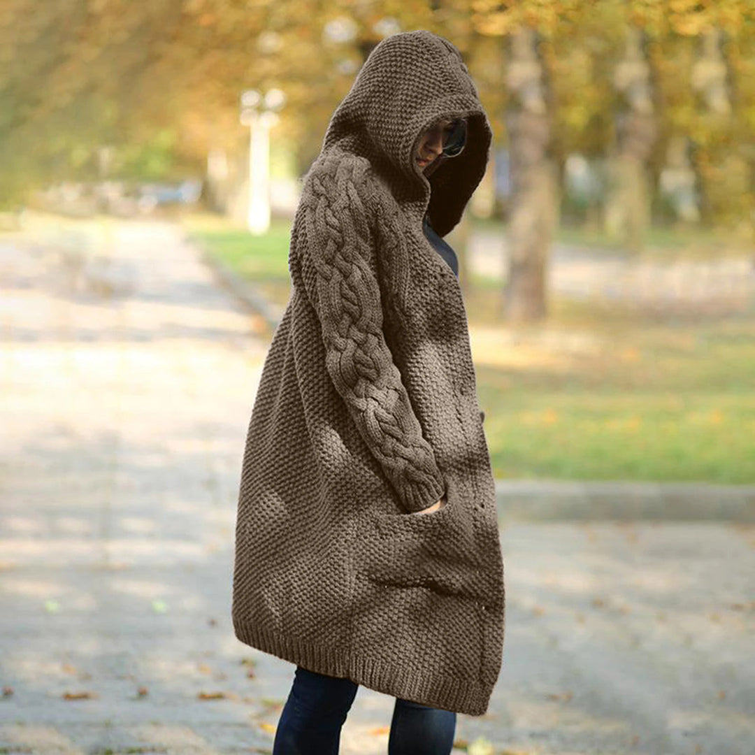 Vrouw in lange, gebreide bruine hoodie met capuchon, herfstbos op achtergrond. Stijlvolle damesmode, warme winterkleding, casual outdoor look.