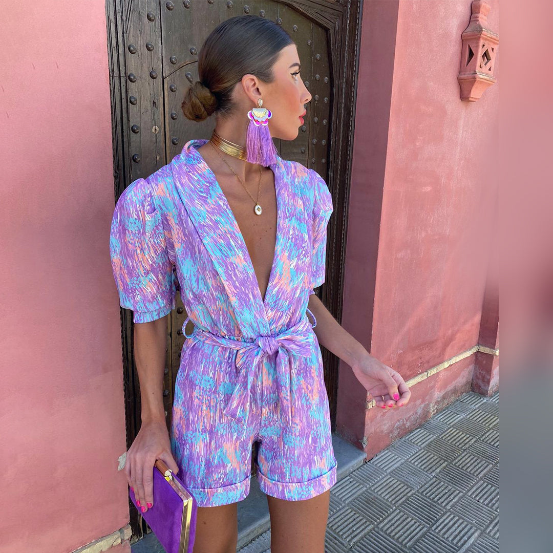 Vrouw in kleurrijke jumpsuit met paarse clutch voor roze muur, modieuze oorbellen en gouden ketting. Zomerse outfit, trendy damesmode.