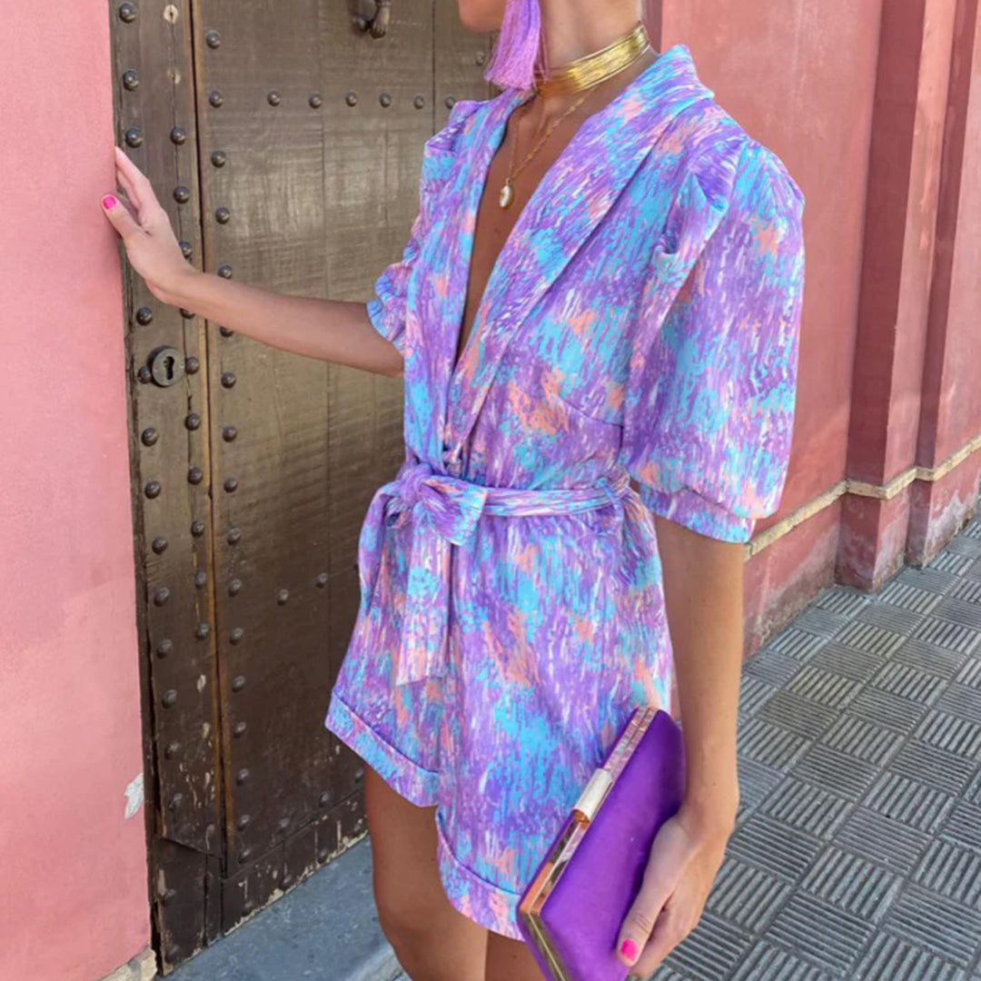 Vrouw in kleurrijke paarse en roze jurk met riem, gouden ketting en paarse clutch, tegen roze muur. Trendy zomeroutfit, modieuze dameskleding.