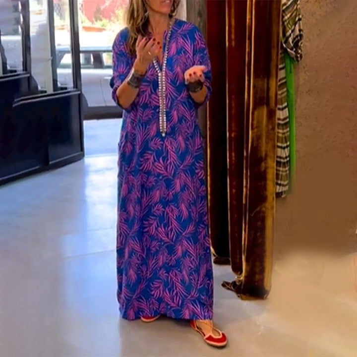 Paarse maxi-jurk met roze bladprint, lange mouwen en V-hals, gedragen door vrouw in moderne winkelomgeving. Trendy damesmode, casual stijl.