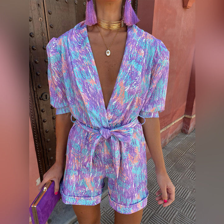 Vrouw in kleurrijke, abstracte paarse jumpsuit met korte mouwen en strikceintuur, houdt een paarse clutch. Trendy zomeroutfit voor dames.