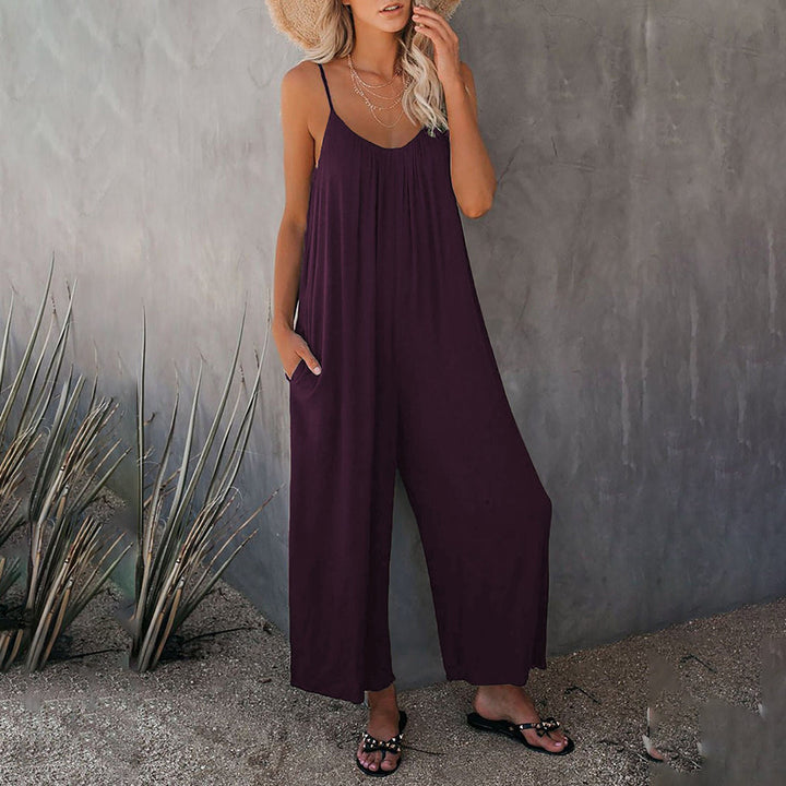 Vrouw in paarse zomerjurk met spaghetti bandjes, strohoed en sandalen, poseert tegen een grijze muur. Perfecte outfit voor casual zomermode.