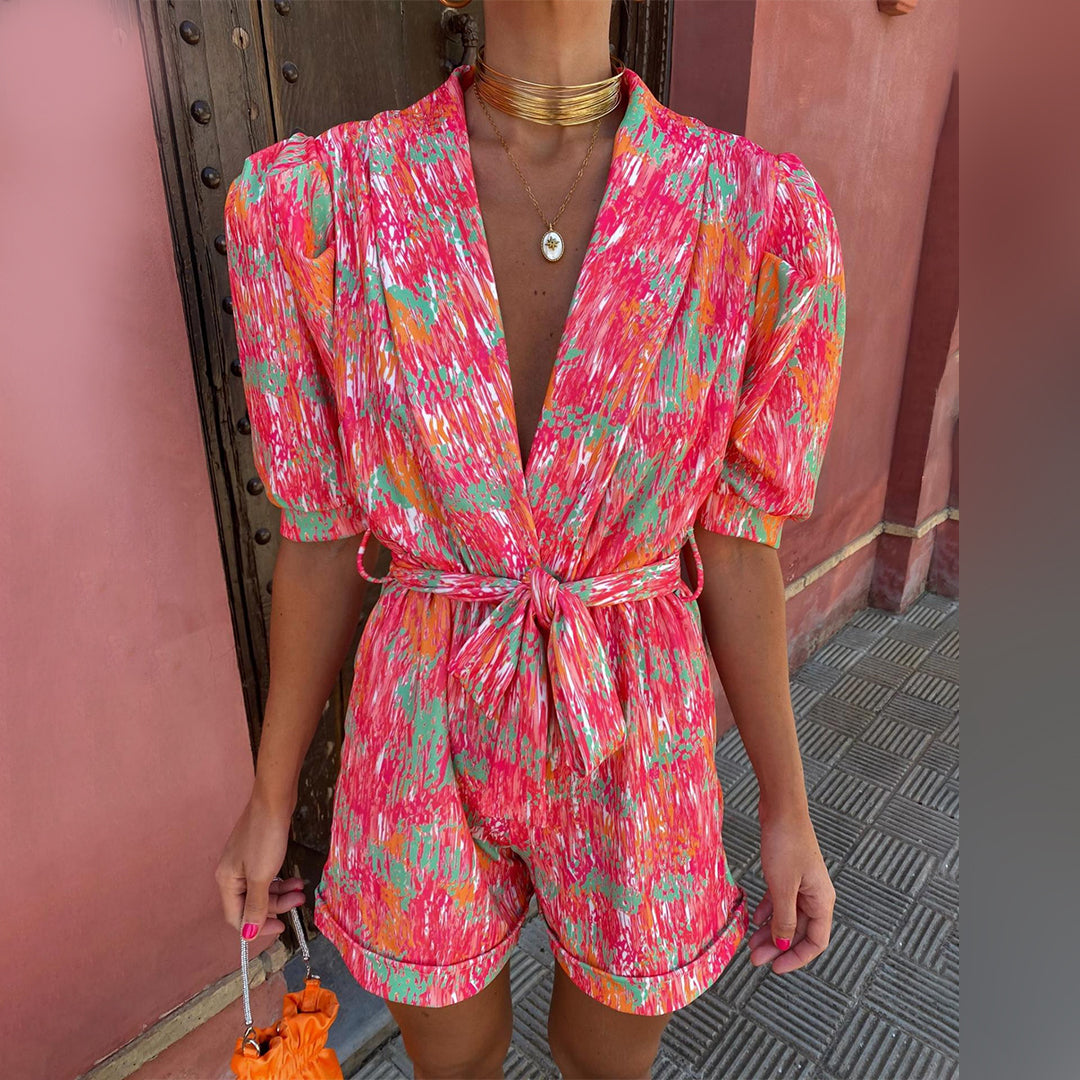 Vrouw in kleurrijke, abstracte print jumpsuit met korte mouwen en strikceintuur, voor houten deur. Trendy zomeroutfit, modieuze dameskleding.