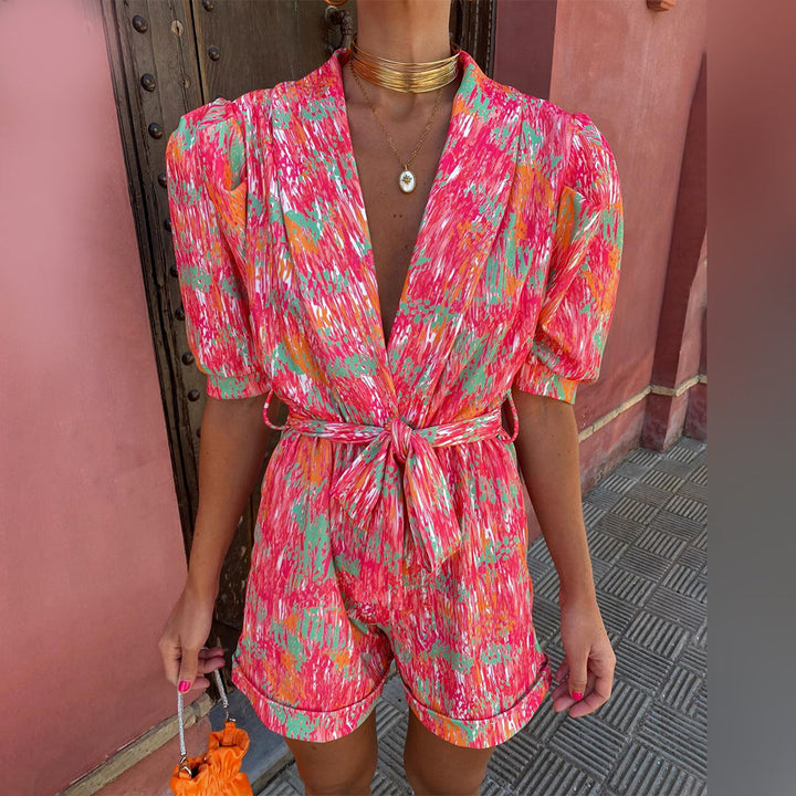 Vrouw in kleurrijke, abstracte print jumpsuit met korte mouwen en strikceintuur, voor houten deur. Trendy zomeroutfit, modieuze dameskleding.