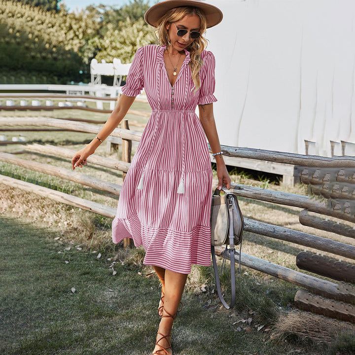 Vrouw in roze gestreepte zomerjurk met hoed en zonnebril, wandelend op gras bij houten hek. Perfecte boho-stijl voor zomermode.