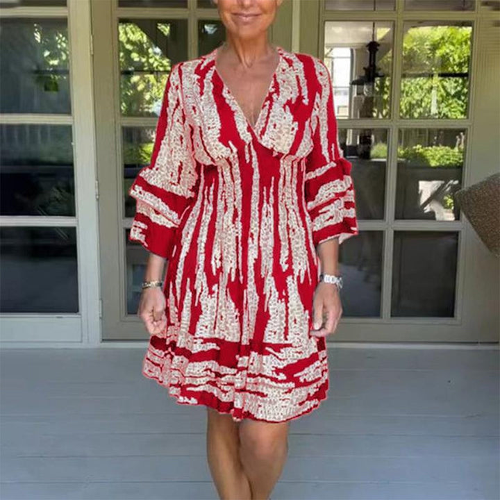 Rode zomerjurk met abstract patroon, driekwart mouwen en V-hals, gedragen door vrouw op veranda. Perfect voor casual en elegante zomerdagen.