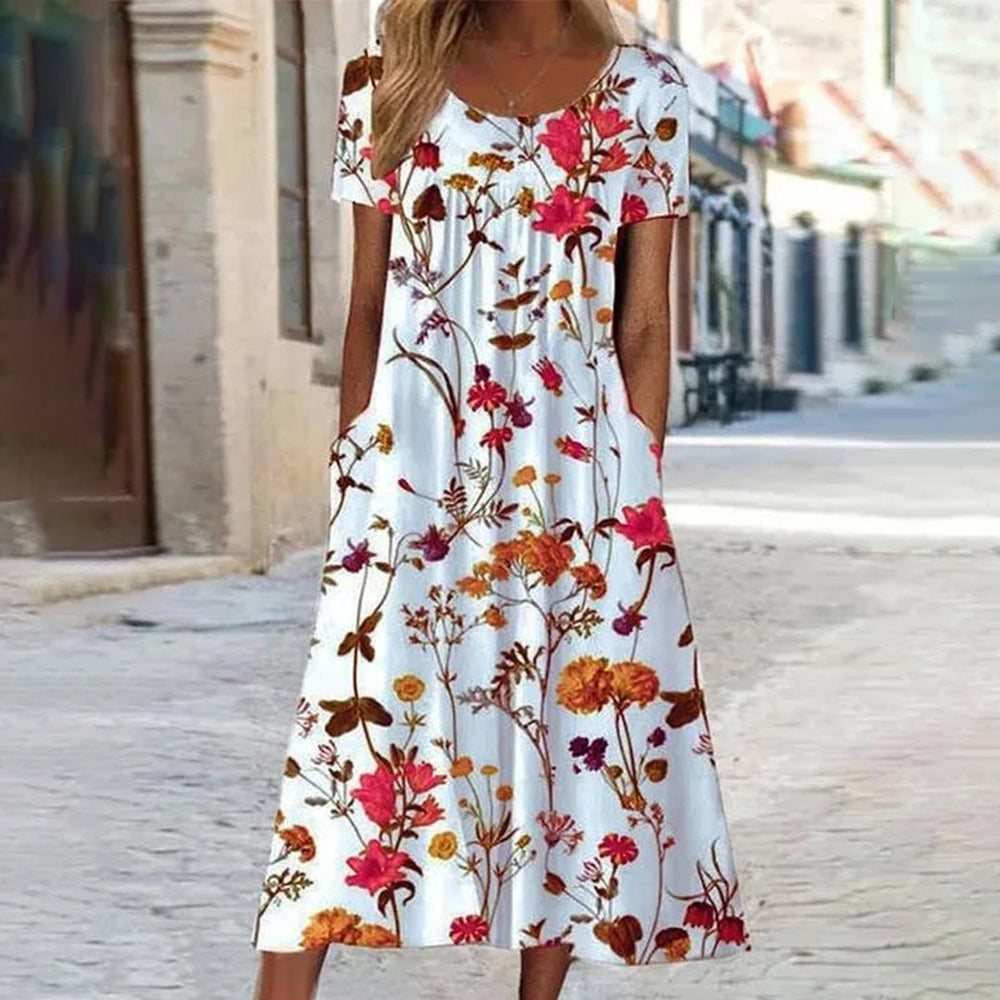 Vrouw in witte zomerjurk met kleurrijke bloemenprint, korte mouwen en zakken, staand op een zonnige straat. Perfect voor casual zomermode.