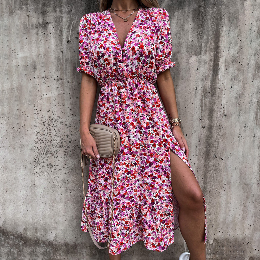 Vrouw in kleurrijke bloemenjurk met split, roze en rode tinten, draagt beige schoudertas. Perfect voor zomermode en casual outfits.