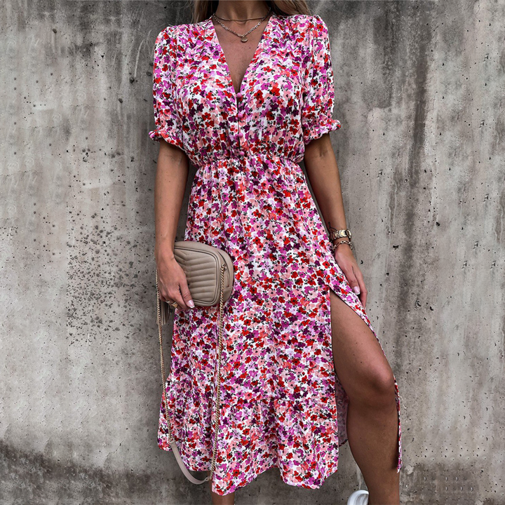 Vrouw in kleurrijke bloemenjurk met split, roze en rode tinten, draagt beige schoudertas. Perfect voor zomermode en casual outfits.