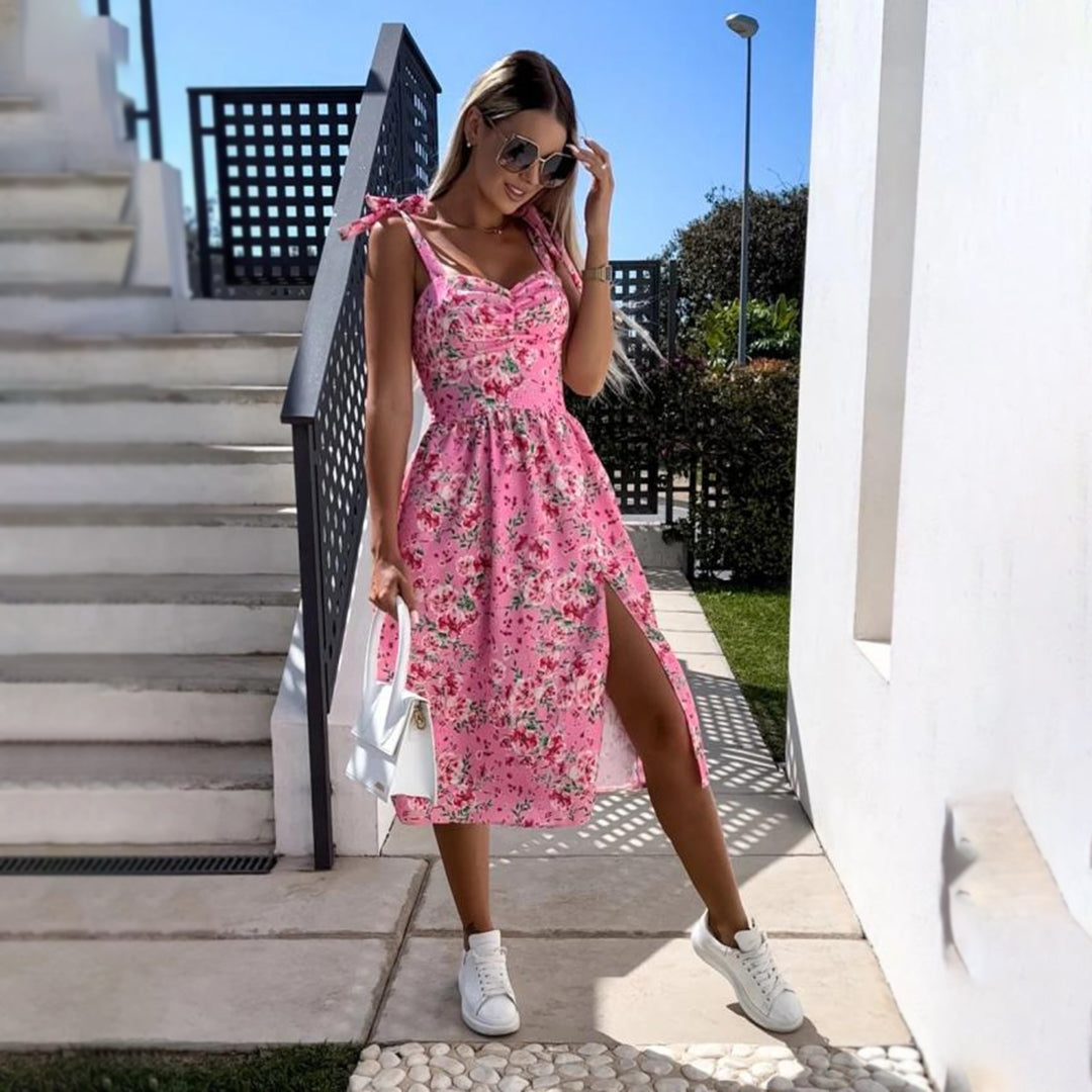 Vrouw in roze bloemenjurk met split, draagt zonnebril en witte sneakers, poseert buiten bij trap. Zomermode, casual stijl, trendy outfit.