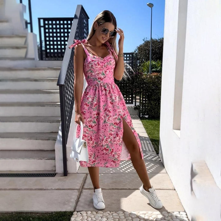 Vrouw in roze bloemenjurk met split, draagt zonnebril en witte sneakers, poseert buiten bij trap. Zomermode, casual stijl, trendy outfit.
