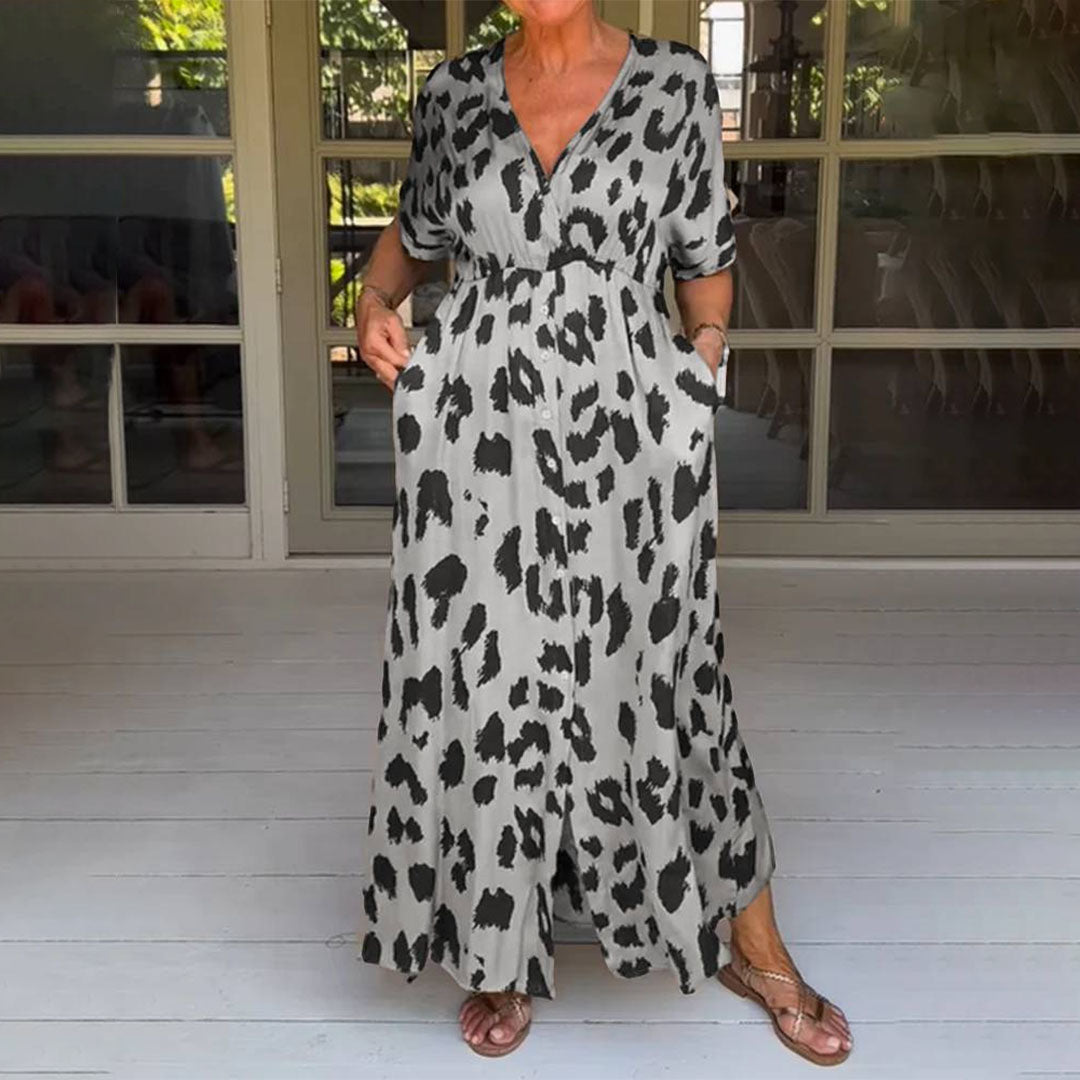 Vrouw in lange, grijze jurk met zwart luipaardprint, korte mouwen en V-hals, staand op een houten vloer voor glazen deuren. Trendy zomerjurk.