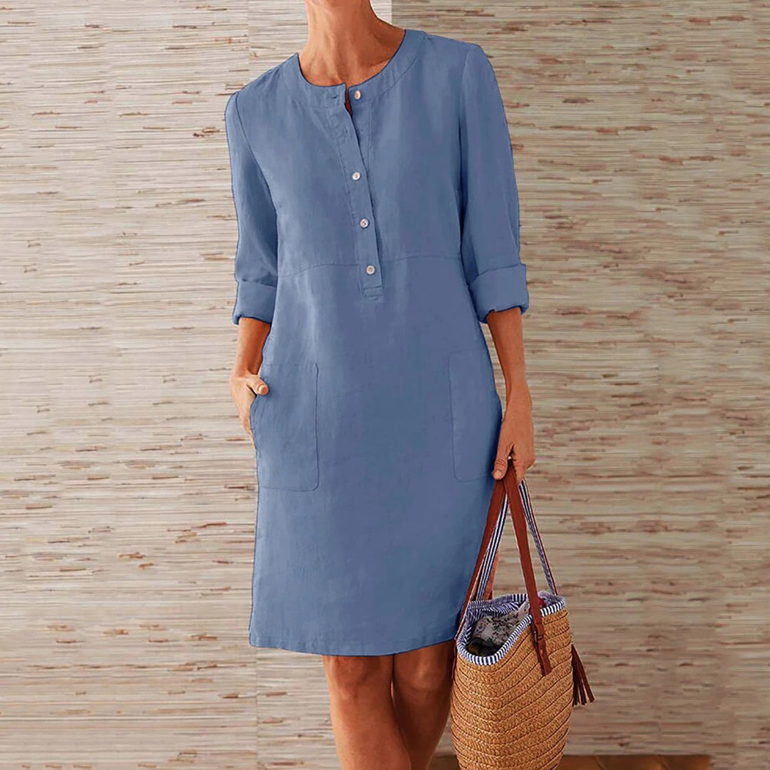 Vrouw in lichtblauwe linnen jurk met knopen, driekwart mouwen en zakken, houdt een strooien tas vast. Perfect voor casual zomeroutfits.