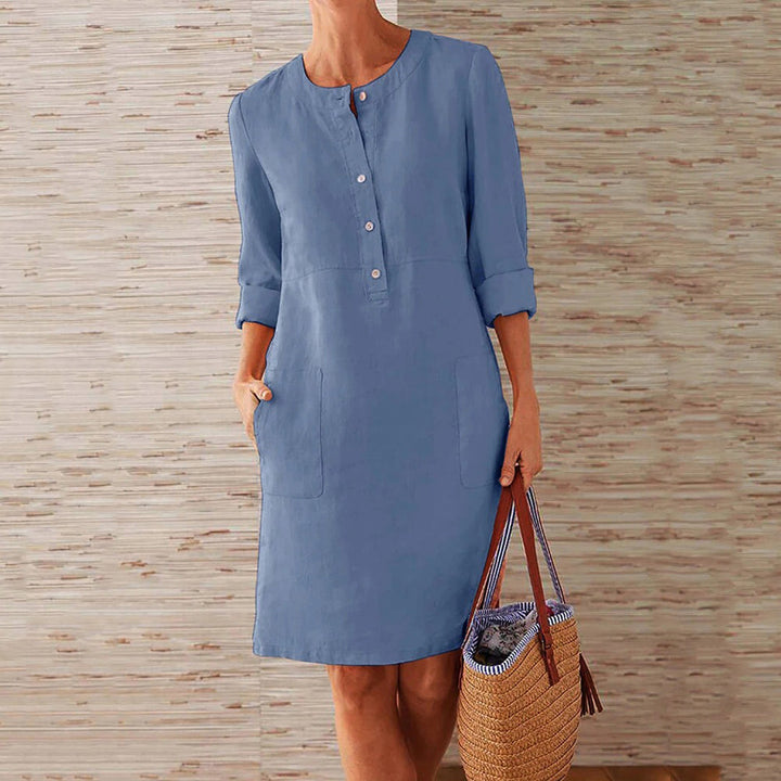 Vrouw in lichtblauwe linnen jurk met knopen, driekwart mouwen en zakken, houdt een strooien tas vast. Perfect voor casual zomeroutfits.