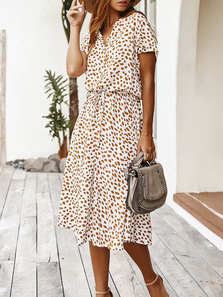 Vrouw in witte zomerjurk met bruine stippen, draagt een grijze handtas en hoed, staand op houten vloer. Perfecte outfit voor casual zomermode.