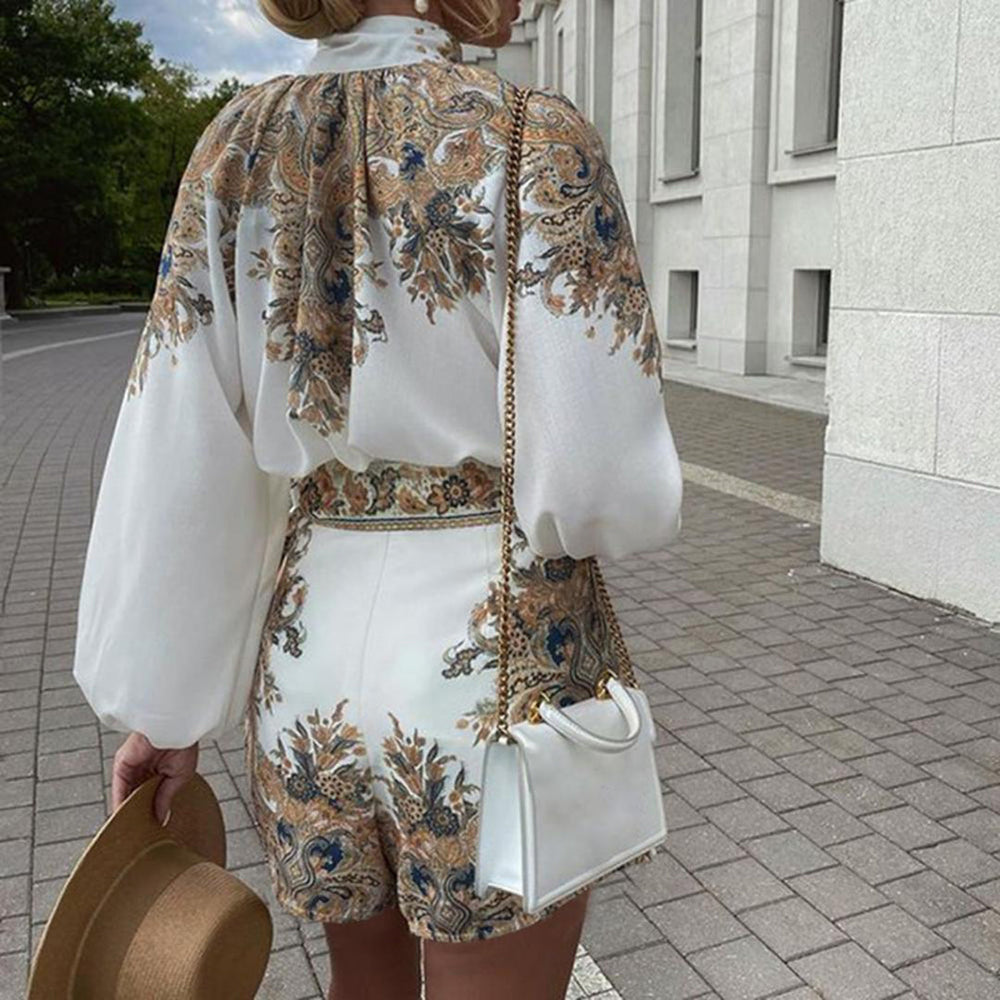 Vrouw in elegante witte jurk met barokke print, draagt een witte handtas en hoed, loopt op een geplaveide straat. Zomeroutfit, modetrend 2023.