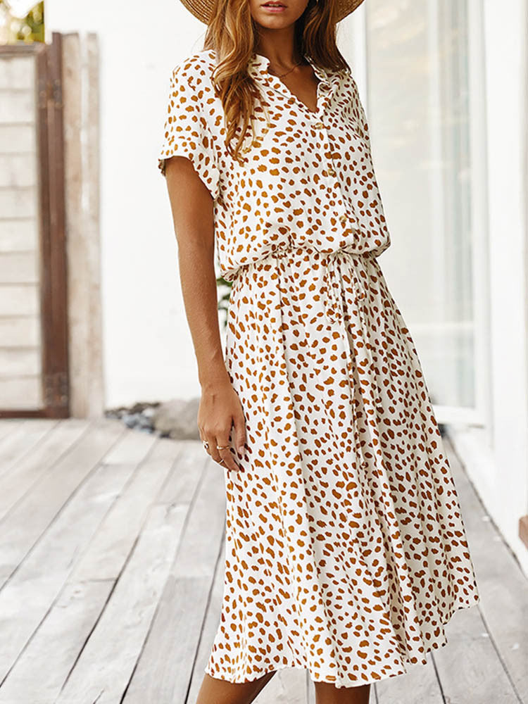 Vrouw in witte zomerjurk met bruine luipaardprint, korte mouwen en V-hals, staand op houten terras. Trendy damesmode, casual stijl.