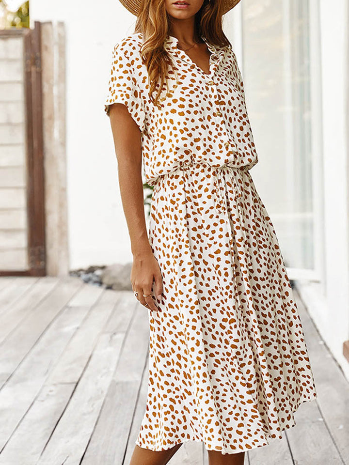 Vrouw in witte zomerjurk met bruine luipaardprint, korte mouwen en V-hals, staand op houten terras. Trendy damesmode, casual stijl.