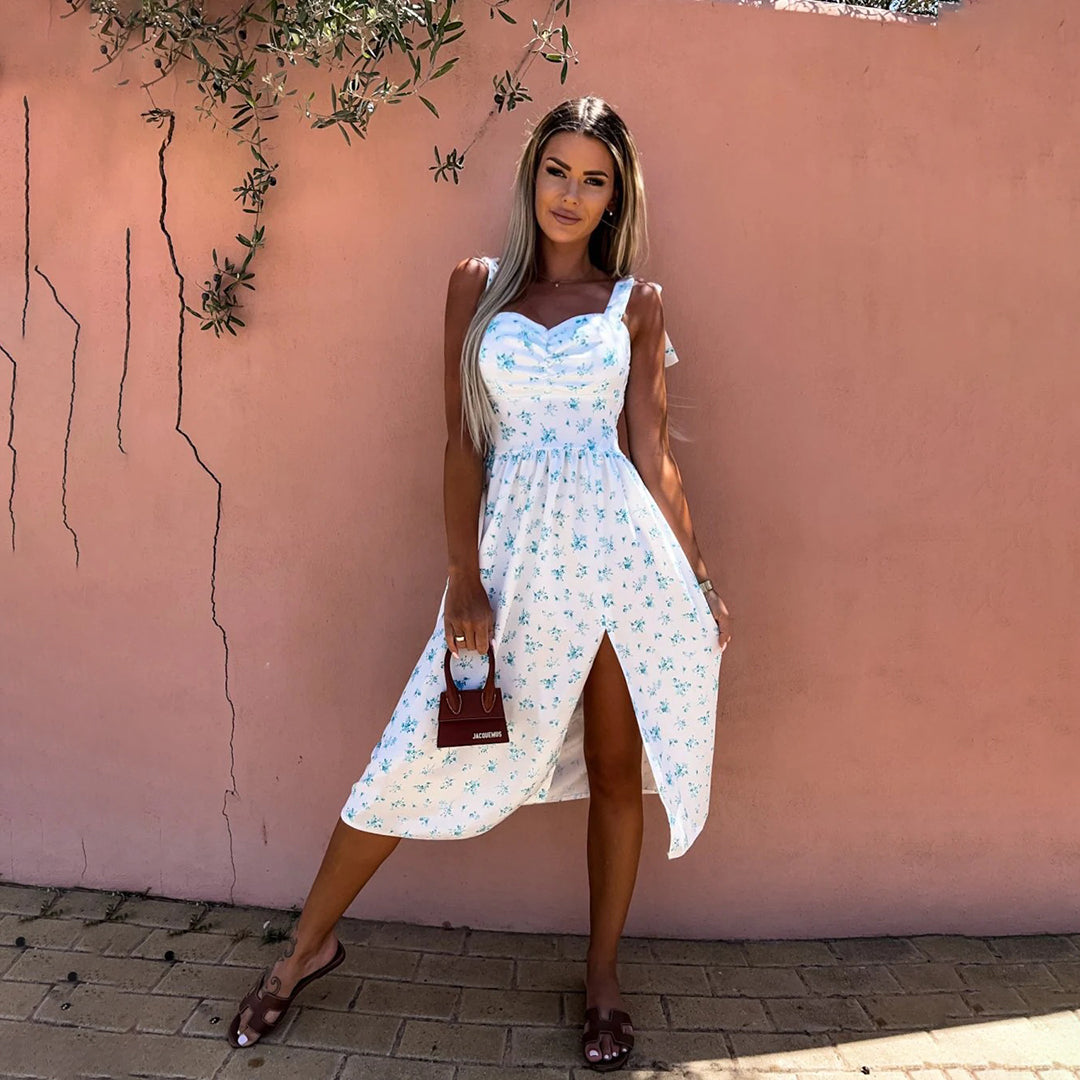Vrouw in witte zomerjurk met blauwe bloemenprint, poseert tegen roze muur. Trendy damesmode, casual zomeroutfit, elegante stijl.