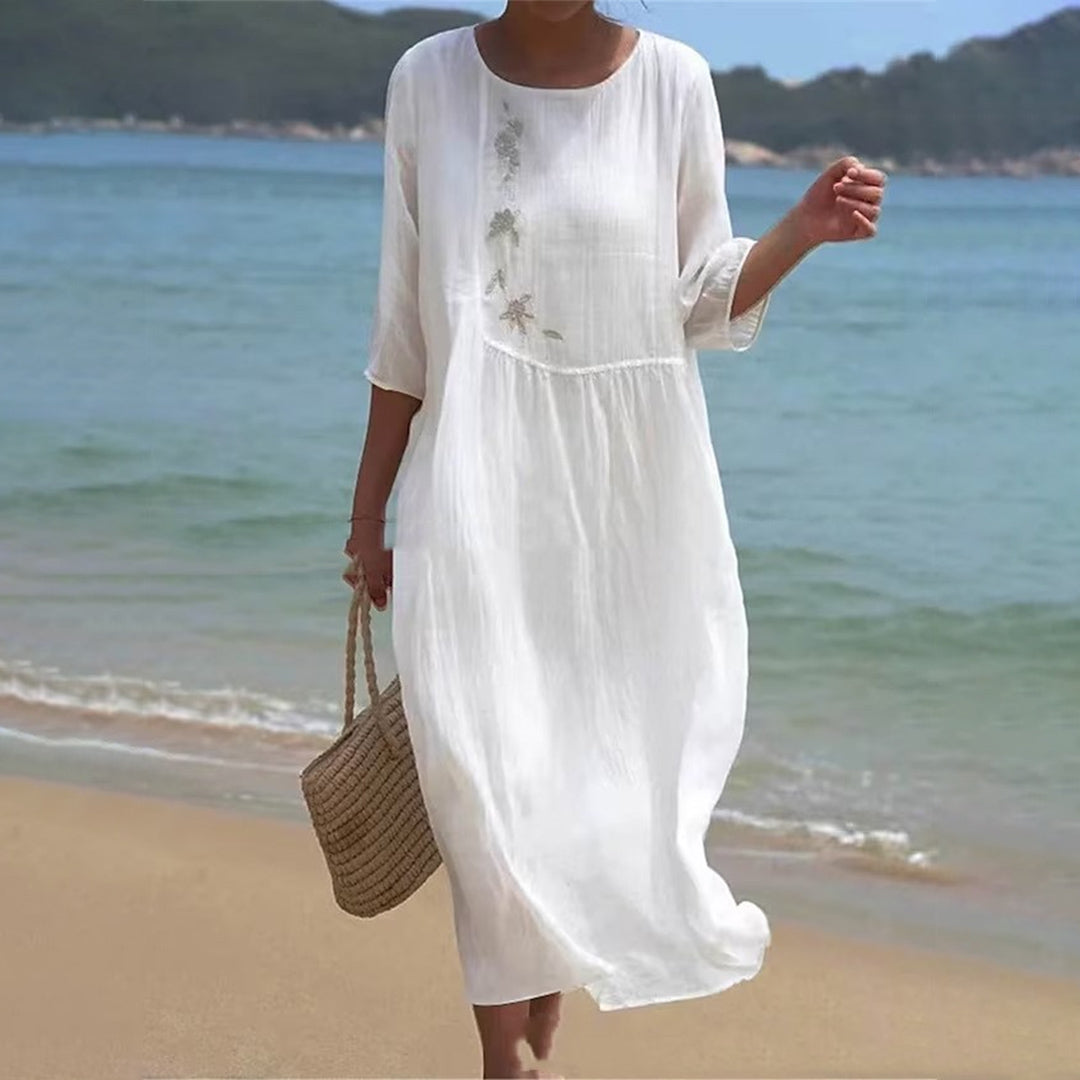 Vrouw in witte zomerjurk met bloemenborduursel, wandelend op strand met rieten tas. Perfect voor strandmode en zomerse stijl.