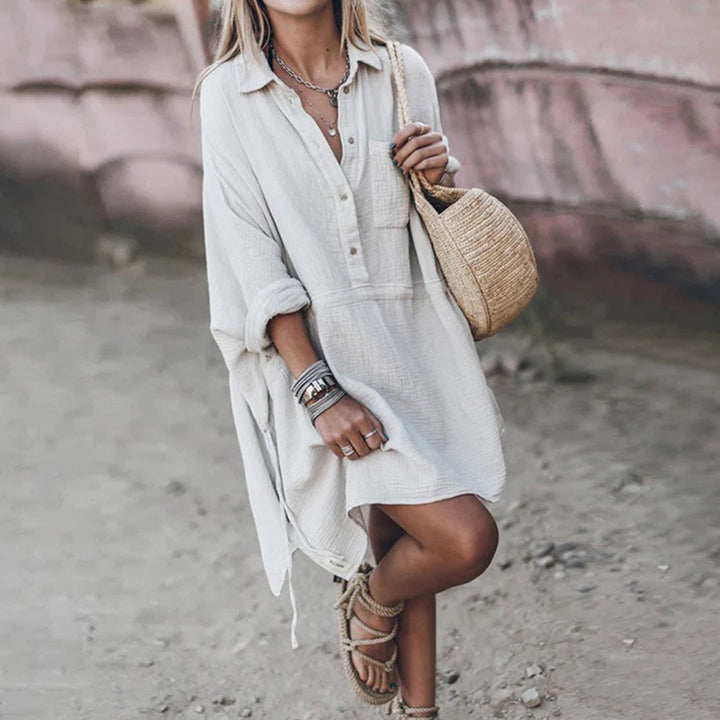 Vrouw in witte, losse linnen jurk met lange mouwen, draagt een strooien tas en sandalen. Boho-stijl, casual zomeroutfit, strandwandeling.