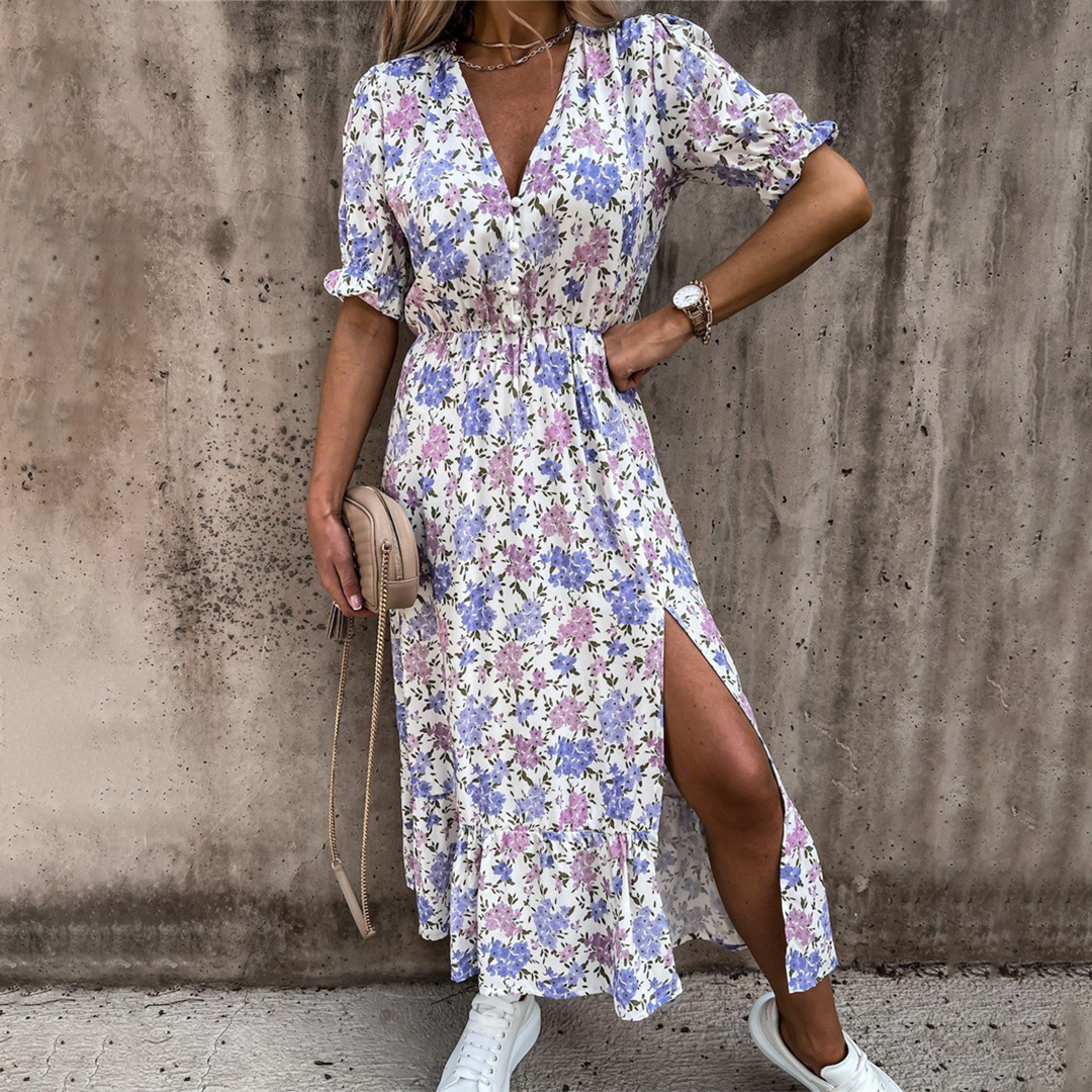 Vrouw in bloemenjurk met paarse en roze print, split aan de zijkant, witte sneakers en beige tas. Perfecte zomeroutfit voor casual gelegenheden.