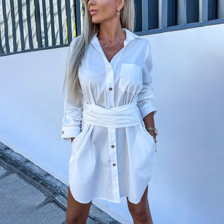 Vrouw in witte overhemdjurk met lange mouwen, knopen en tailleband, buiten bij moderne muur. Perfect voor casual en stijlvolle zomermode.