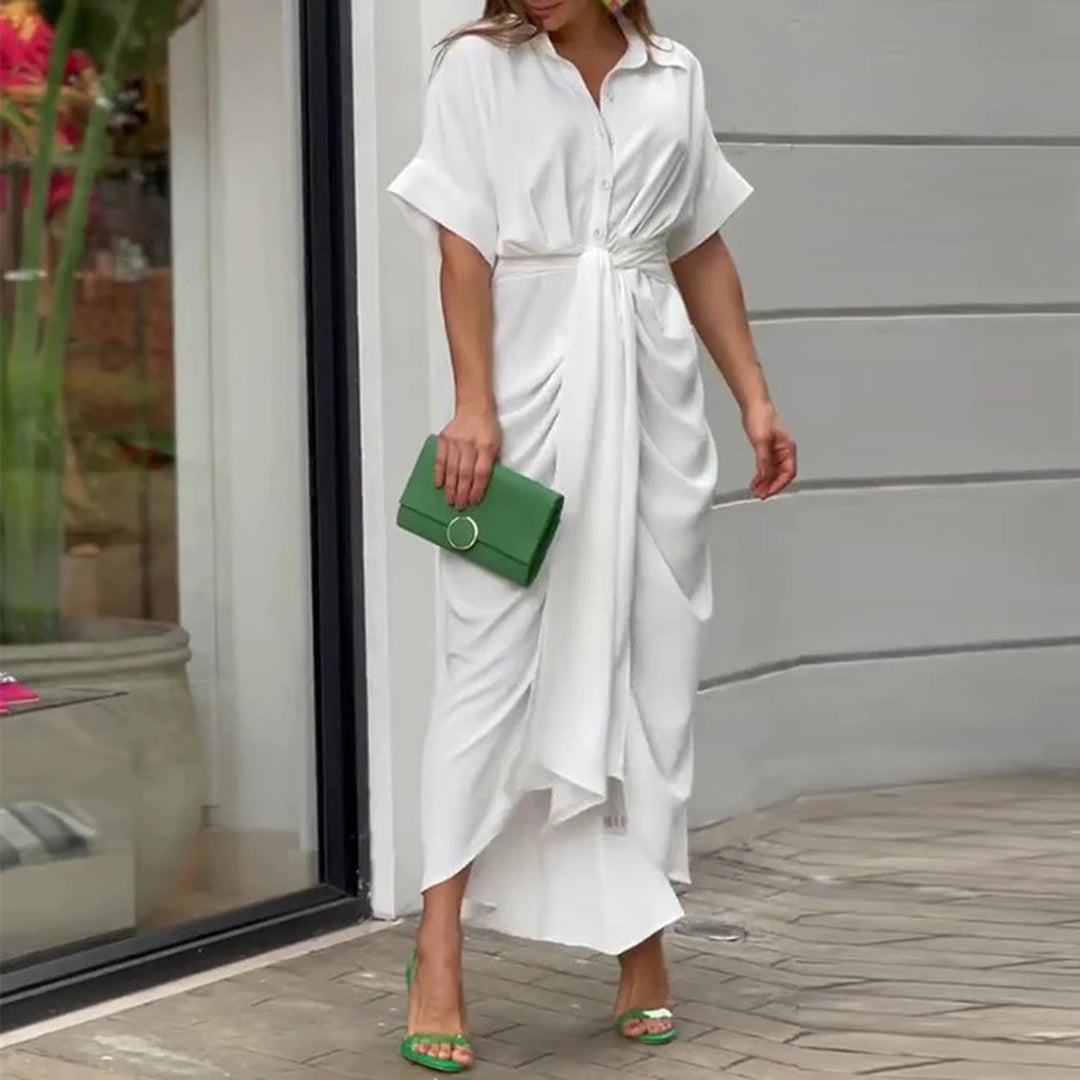 Witte maxi-jurk met knoopdetail, gedragen door vrouw met groene clutch en sandalen. Stijlvolle zomermode, perfect voor elegante gelegenheden.