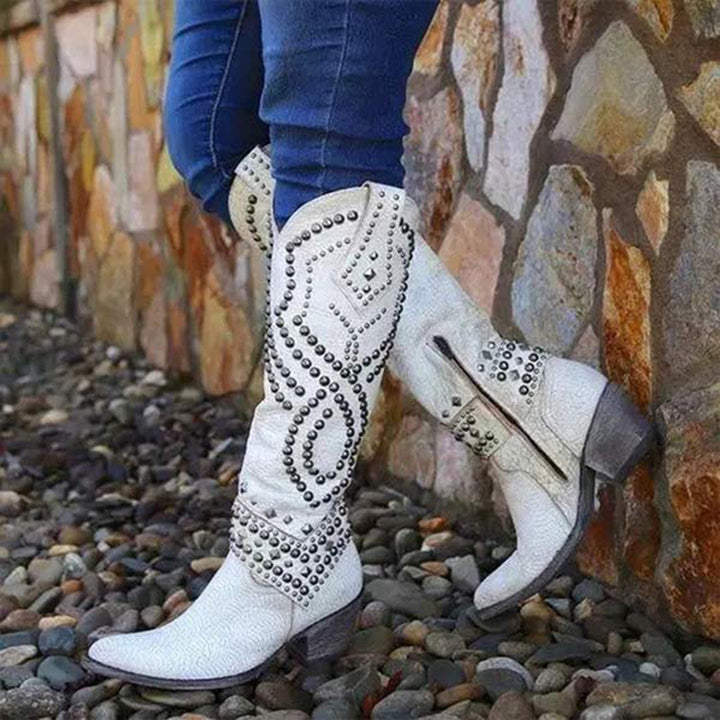 Witte cowboylaarzen met studs, gedragen door persoon in blauwe jeans, tegen stenen muur. Trendy westernstijl, damesmode, herfstaccessoire.