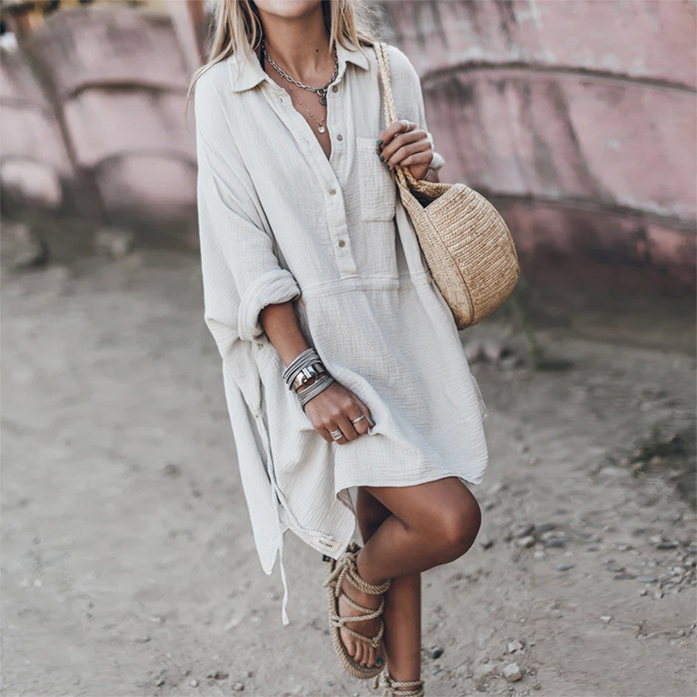 Vrouw in witte, losse zomerjurk met lange mouwen, draagt een strooien tas en sandalen. Perfecte boho-stijl voor strandvakanties en zomermode.