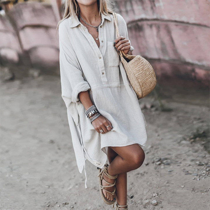 Vrouw in witte, losse zomerjurk met lange mouwen, draagt een strooien tas en sandalen. Perfecte boho-stijl voor strandvakanties en zomermode.