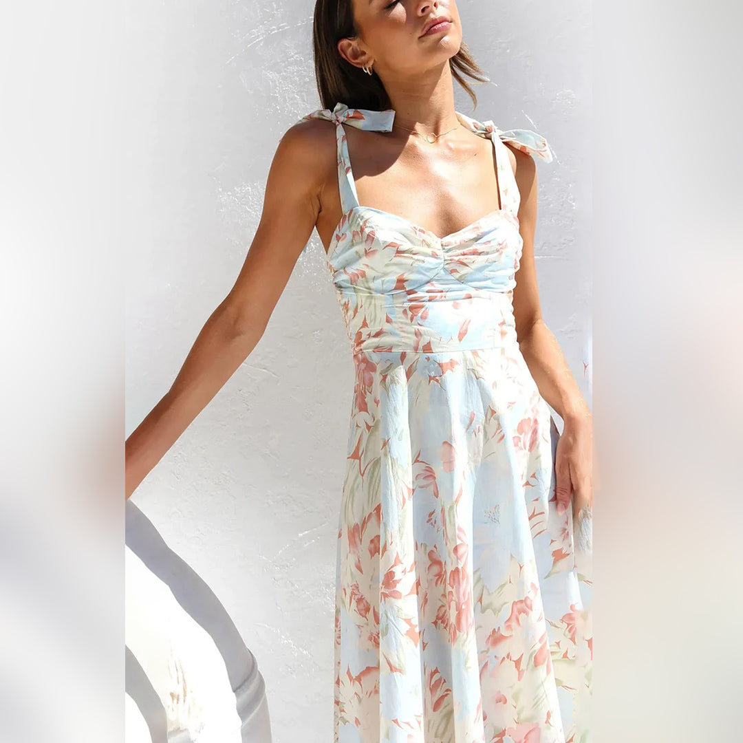 Vrouw in zomerse bloemenjurk met strikbandjes, pastelkleurig met abrikoos en blauw patroon, tegen witte achtergrond. Perfect voor lente- en zomertrends.