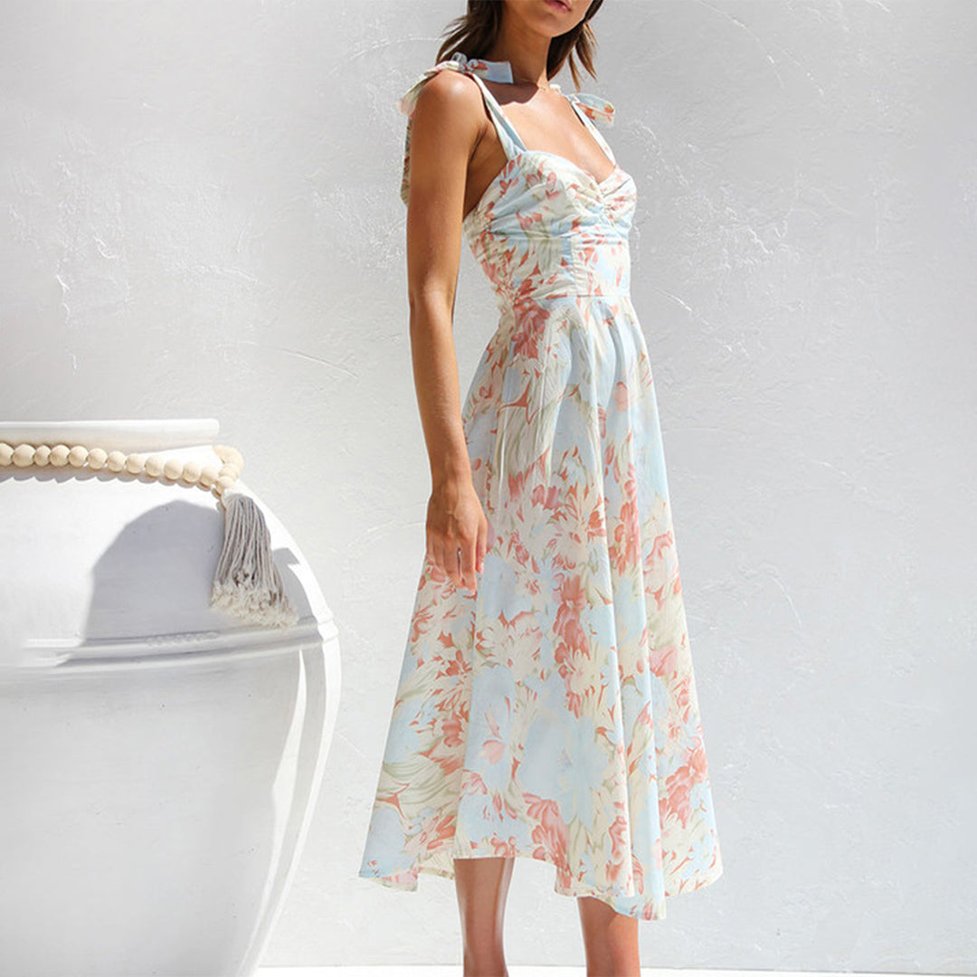 Vrouw in zomerse bloemenjurk met pastelkleurige print, staand naast grote witte vaas. Perfect voor lente- en zomermode.
