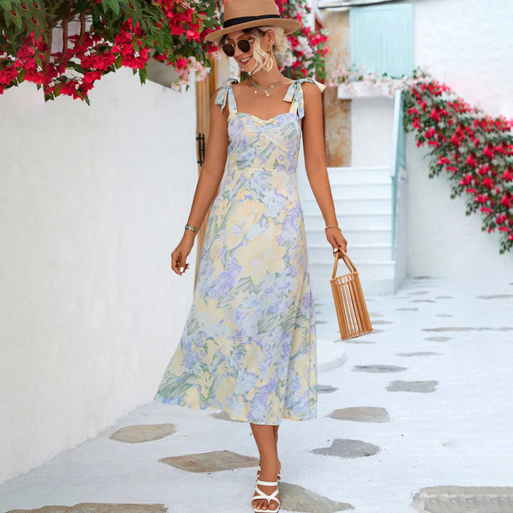 Vrouw in zomerse bloemenjurk met hoed en bamboe tas, wandelend langs witte muur met bloemen. Perfecte outfit voor zomermode en strandvakantie.