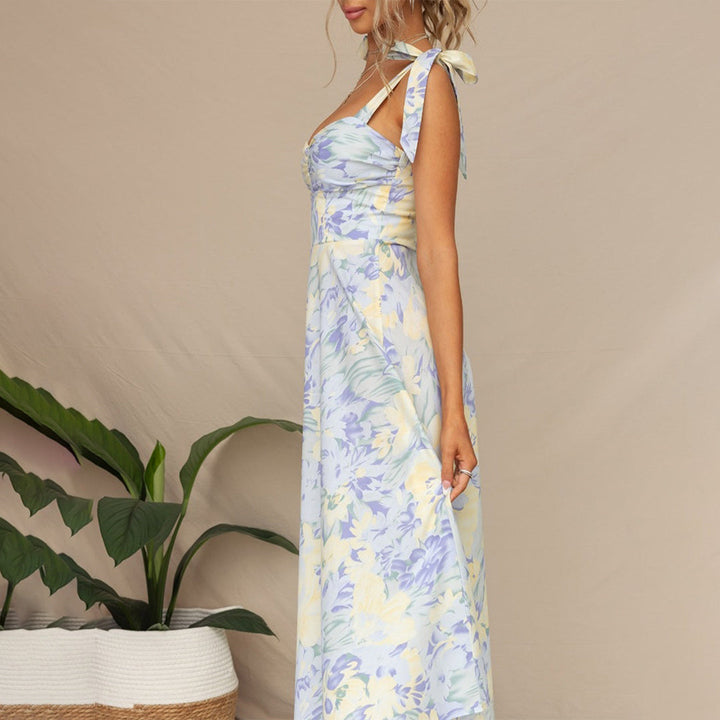 Vrouw in lange, blauwe bloemenjurk met strikbandjes, staand naast groene plant. Zomerse damesmode, elegante maxi-jurk, casual stijl.