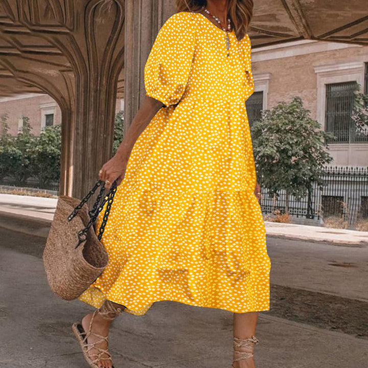 Vrouw in gele jurk met witte stippen, draagt een rieten tas, loopt onder een overdekte doorgang. Zomerse mode, casual stijl, stedelijke achtergrond.