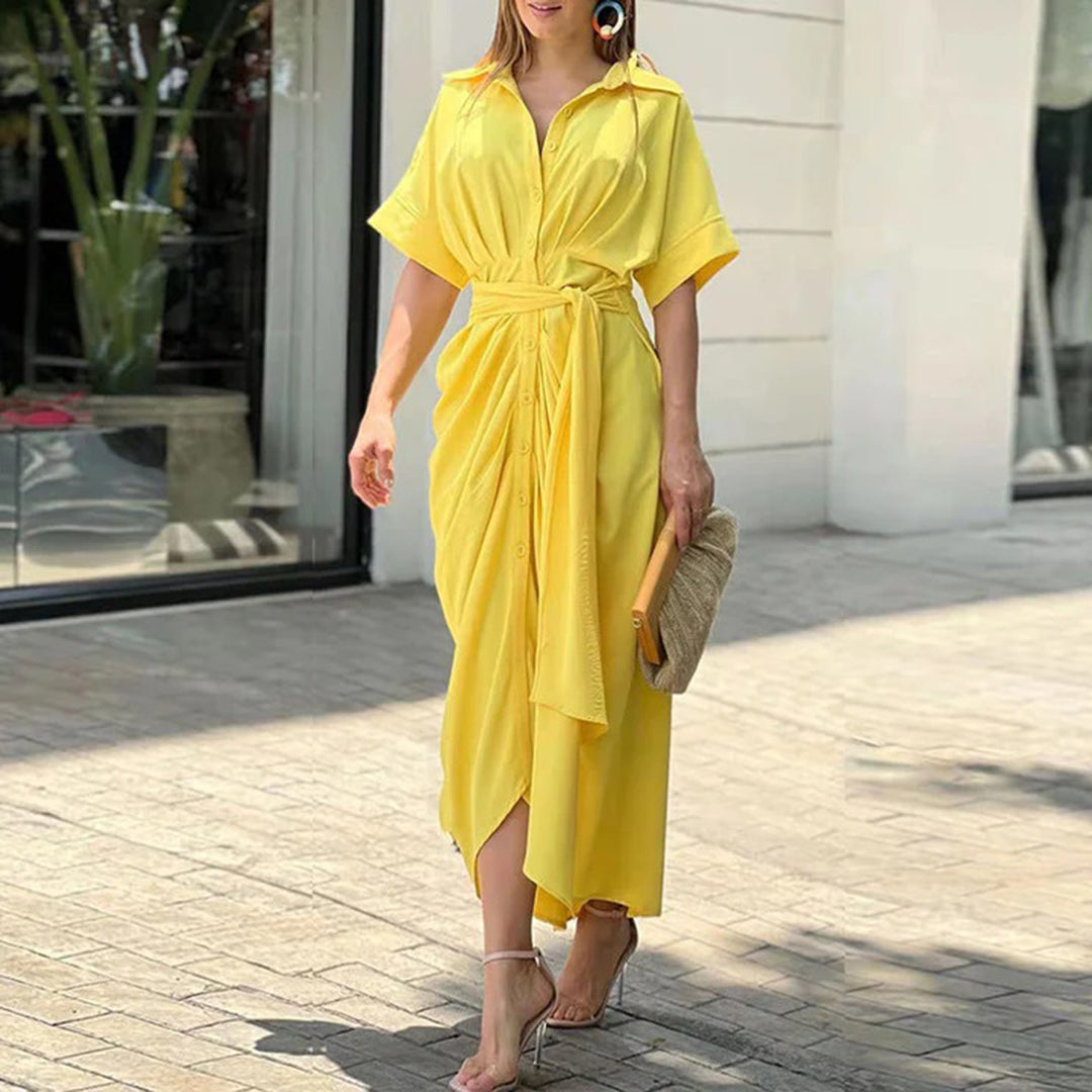 Vrouw in gele zomerjurk met knoopdetail, draagt een stro clutch en sandalen, loopt buiten bij een moderne winkel. Perfecte zomermode.