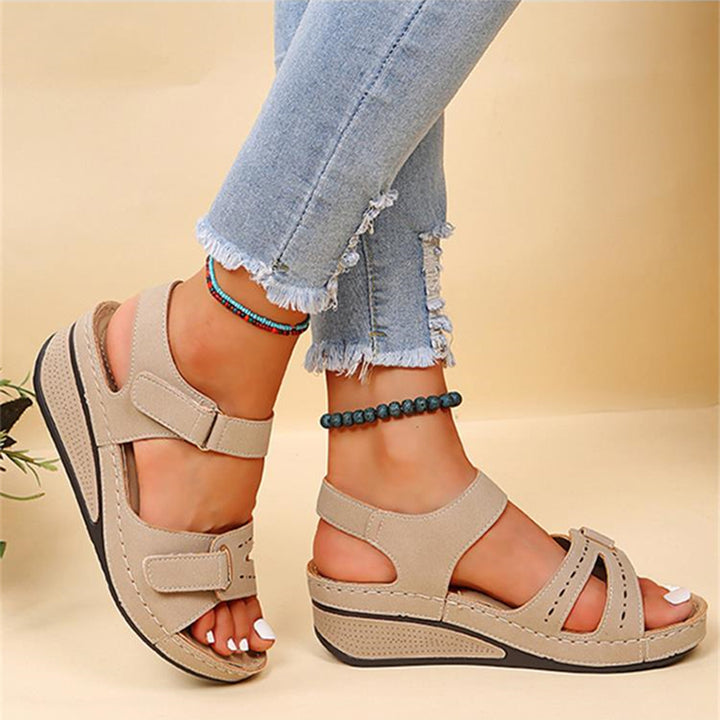 Beige sandalen met klittenband, gedragen door een persoon met blauwe jeans en enkelarmbanden. Comfortabele zomerschoenen voor dames.